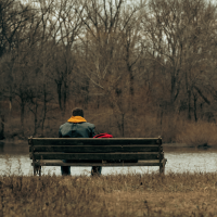 A man is sat on a bench beside a river. It's a grey, autumnal scene, with bare trees.
