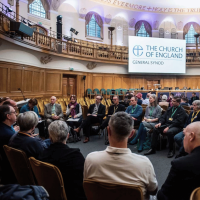 A group of people are sat on chairs in a circle, in the main hall of Church House.
