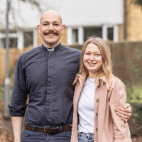 Nik is wearing a grey shirt and a white clerical collar. Heidi is wearing a white t-shirt and a salmon jacket. They are stood outside, and Nik has his arm around Heidi.