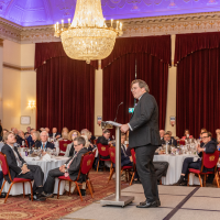 The Revd Ben Cahill-Nicholls is dressed in a black suit and standing behind a podium. He is addressing a room of seated guests. A grand, bright chandelier is hanging from the ceiling.