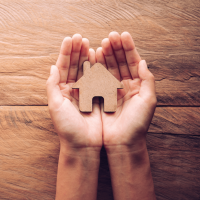 On a wooden table, a pair of outstretched, open hands are holding a small wooden icon of a house.
