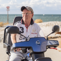 Martin is sat on a dark blue mobility scooter. Behind him is a long stretch of sandy beach, leading to the ocean.