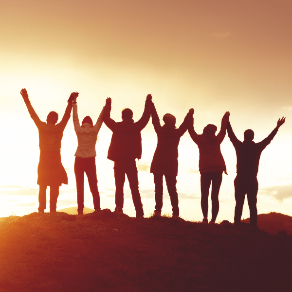 Six people joyously holding raised hands, silhouetted against a sunset, on top of a mountain